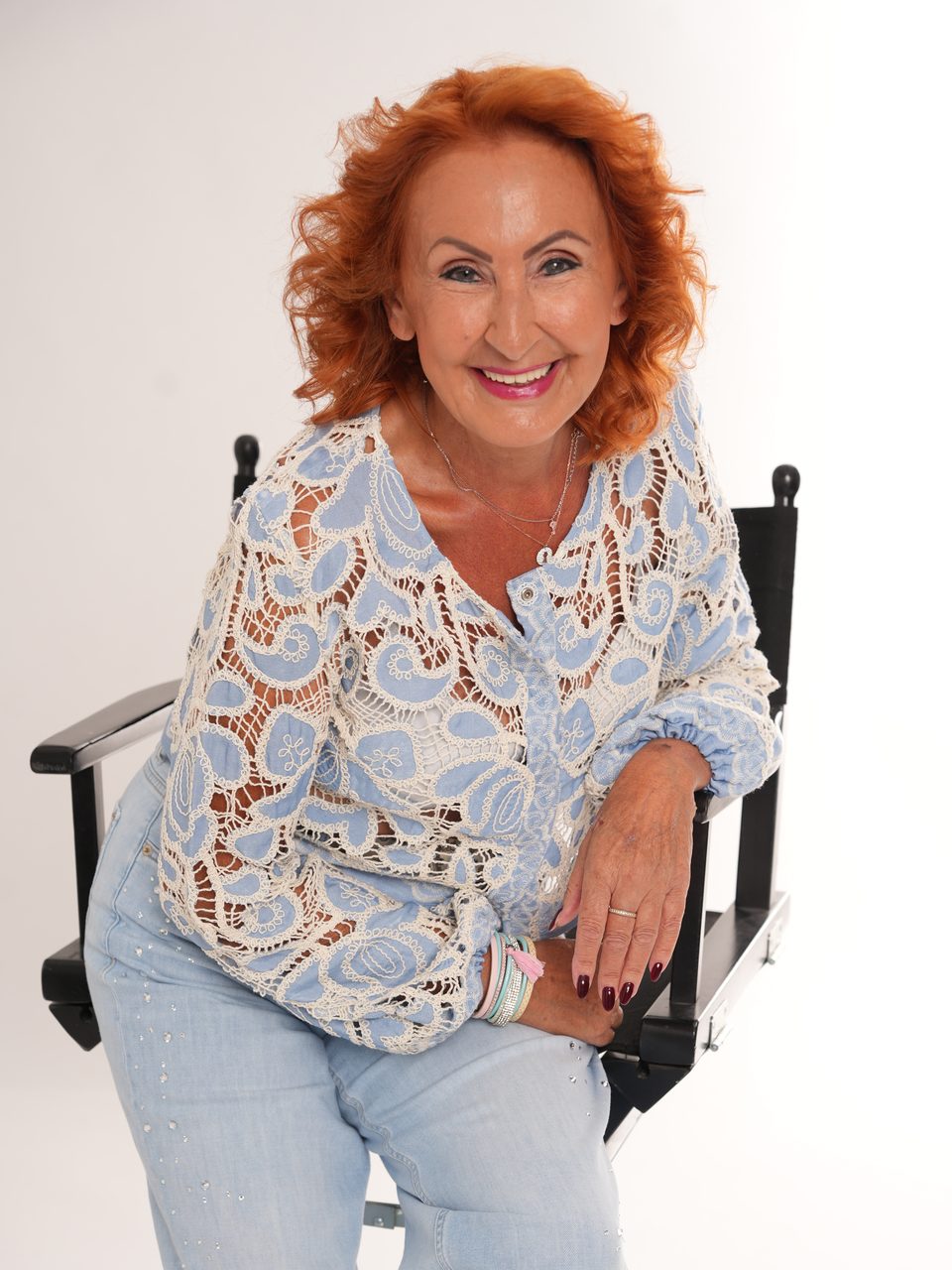 Marietta in hellblauer Spitzenbluse und Jeans auf einem Regiestuhl, lachend